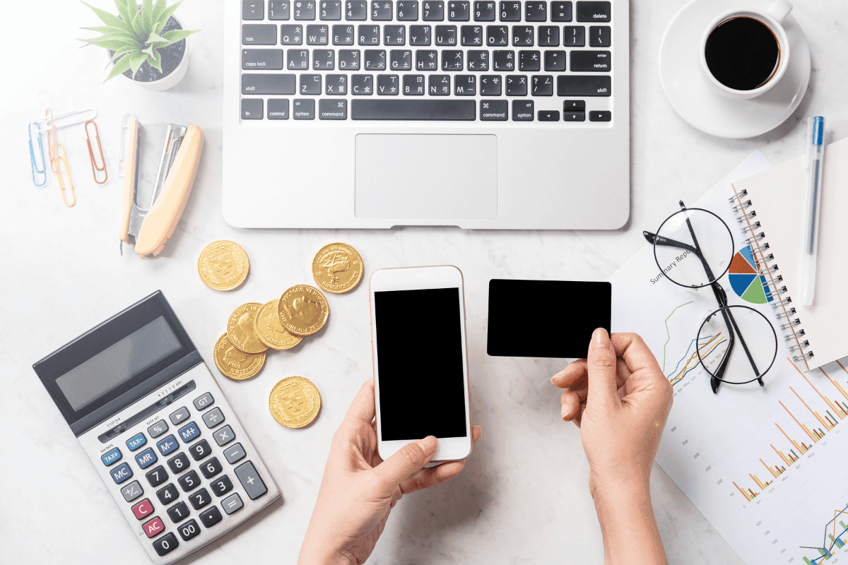 Eine Person sitzt vor ihrem Laptop und hat ihr Handy in der einen sowie eine Kreditkarte in der anderen Hand. Daneben liegen Münzen sowie ein Taschenrechner, eine Brille und mehrere Kalkulationsgrafik.