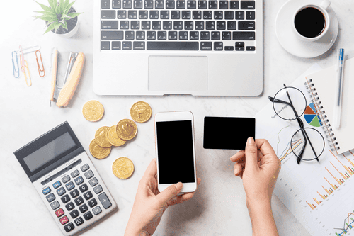 Eine Person sitzt vor ihrem Laptop und hat ihr Handy in der einen sowie eine Kreditkarte in der anderen Hand. Daneben liegen Münzen sowie ein Taschenrechner, eine Brille und mehrere Kalkulationsgrafik.