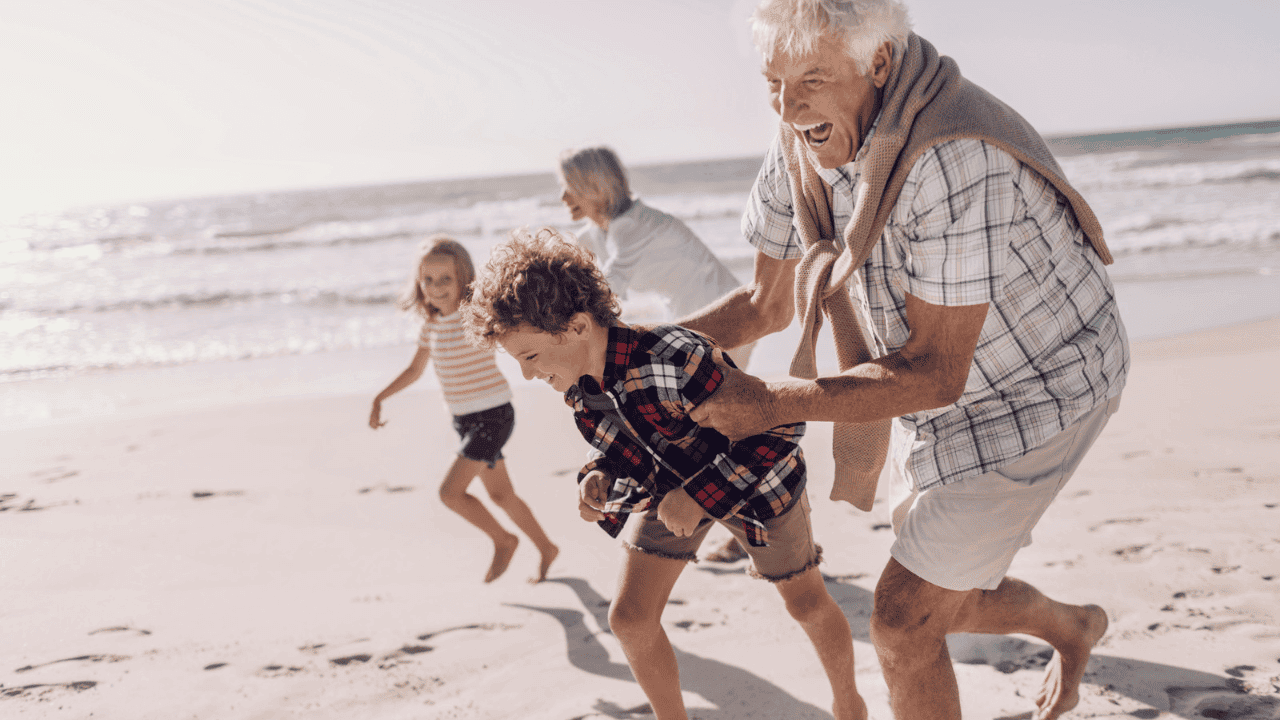 Oma und Opa toben mit Enkel und Enkelin auf einem weißen Sandstrand.