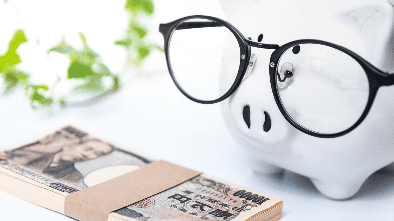 Ein kreatives Bild, das ein Sparschwein mit einer schwarzen Brille darstellt, neben einem Bündel japanischer Yen-Noten. Im Hintergrund ist unscharf grünes Laub zu sehen, was auf Wachstum und finanzielle Planung anspielt.