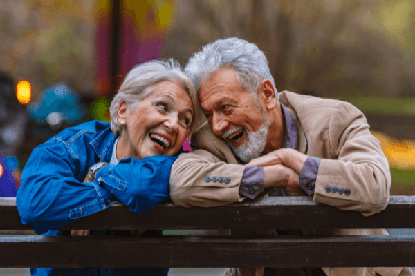 Zwei ältere Menschen, die sichtlich Freude und Zuneigung teilen, lehnen sich eng aneinander auf einer Parkbank. Sie blicken sich liebevoll an, umgeben von herbstlichen Farben, ein Bild des Glücks und der Zufriedenheit im Ruhestand.