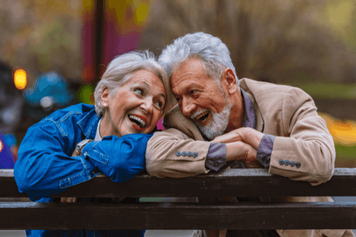 Zwei ältere Menschen, die sichtlich Freude und Zuneigung teilen, lehnen sich eng aneinander auf einer Parkbank. Sie blicken sich liebevoll an, umgeben von herbstlichen Farben, ein Bild des Glücks und der Zufriedenheit im Ruhestand.