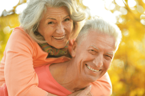 Ein glückliches älteres Paar genießt ihre gemeinsame Zeit im goldenen Herbstlicht. Der Mann trägt ein begeistertes Lächeln, während die Frau mit grauen Haaren fröhlich auf seinen Schultern sitzt, symbolisch für einen sorgenfreien Ruhestand.