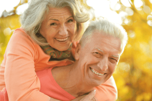 Ein glückliches älteres Paar genießt ihre gemeinsame Zeit im goldenen Herbstlicht. Der Mann trägt ein begeistertes Lächeln, während die Frau mit grauen Haaren fröhlich auf seinen Schultern sitzt, symbolisch für einen sorgenfreien Ruhestand.