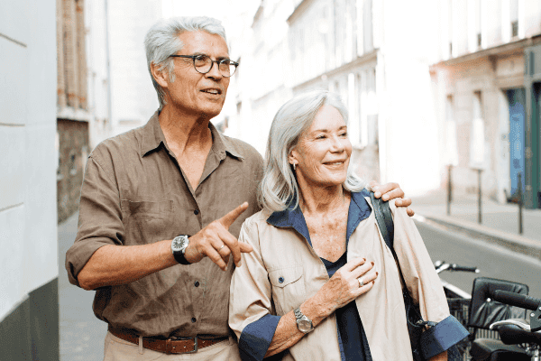 Ein strahlendes älteres Paar, das gemeinsam durch eine urbane Gasse spaziert, symbolisiert die freudige Lebensphase im Ruhestand. Der Mann, lässig gekleidet in einem hellen Hemd und dunkler Hose, zeigt begeistert auf etwas außerhalb des Bildes, während die Frau, ebenfalls entspannt gekleidet mit einer hellen Jacke über den Schultern, ihn mit einem liebevollen Lächeln ansieht. Dieses Bild repräsentiert ein sorgloses Rentnerleben, welches durch eine fundierte Ruhestandsplanung ermöglicht wurde.