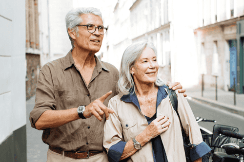 Ein strahlendes älteres Paar, das gemeinsam durch eine urbane Gasse spaziert, symbolisiert die freudige Lebensphase im Ruhestand. Der Mann, lässig gekleidet in einem hellen Hemd und dunkler Hose, zeigt begeistert auf etwas außerhalb des Bildes, während die Frau, ebenfalls entspannt gekleidet mit einer hellen Jacke über den Schultern, ihn mit einem liebevollen Lächeln ansieht. Dieses Bild repräsentiert ein sorgloses Rentnerleben, welches durch eine fundierte Ruhestandsplanung ermöglicht wurde.