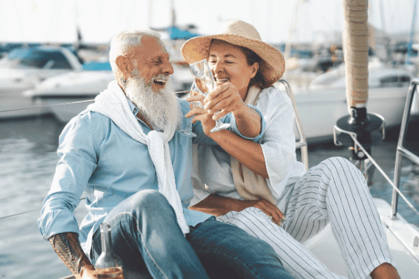 Ein glückliches, älteres Paar genießt gemeinsame Zeit auf einer Yacht. Der Mann mit vollem, weißem Bart und die Frau mit einem Strohhut stoßen mit Weingläsern an. Sie tragen sommerliche Kleidung und lachen miteinander, während sie in die Kamera schauen. Der Hintergrund zeigt ruhiges Hafenwasser mit weiteren Booten, was ein Gefühl von Freiheit und sorglosem Genuss im Ruhestand vermittelt. Das Bild strahlt Wärme, Liebe und die Früchte langfristiger finanzieller Planung aus.