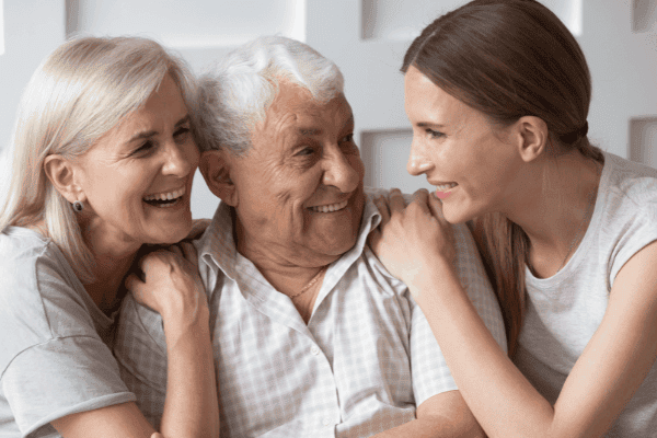 Ein herzliches Familienbild, das eine ältere Dame und einen Herrn zeigt, beide lächelnd und eng umarmt von einer jüngeren Frau, wahrscheinlich ihrer Tochter. Alle drei tragen fröhliche Gesichter und weiße Oberteile. Dieses Bild fängt einen Moment des Glücks und der familiären Bindung ein, der die Bedeutung des emotionalen Werts einer sorgfältigen Vermögensübertragung und Nachlassplanung unterstreicht.