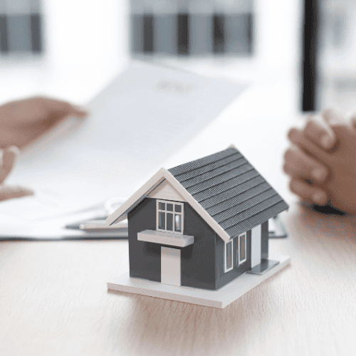Das Bild zeigt eine Beratungssituation, in der eine Person ein Dokument über den Tisch zu einer anderen Person reicht, die erwartungsvoll die Hände verschränkt hält. Im Vordergrund ist ein kleines Modellhaus auf dem Tisch platziert, was den Fokus auf Immobilieninvestitionen und Beratungsdienstleistungen legt. Die Szene spielt sich in einem hellen Büro ab und vermittelt ein professionelles, kundenorientiertes Beratungsgespräch. Ideal für eine Webseite, die Dienstleistungen rund um Immobilien und Finanzberatung anbietet.