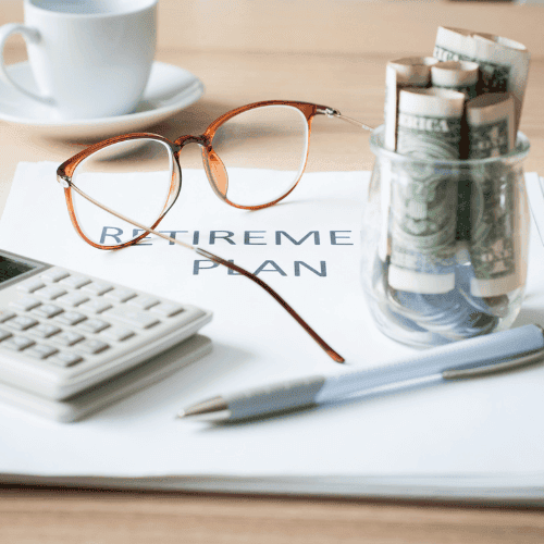 Arbeitsplatz mit Fokus auf Altersvorsorgeplanung, bestehend aus einem Taschenrechner, einer Brille und einem Kugelschreiber auf einem Stapel weißer Blätter mit der Überschrift 'RETIREMENT PLAN'. Neben den Unterlagen steht ein Glas mit gerollten Geldscheinen, im Hintergrund eine Kaffeetasse auf einem Holztisch.