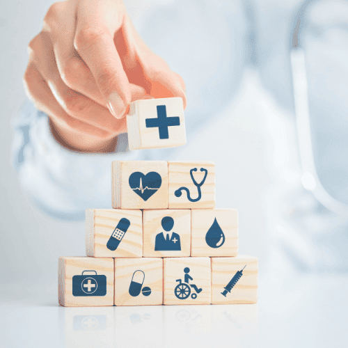 Ein konzeptionelles Bild, das eine Gesundheitsberatung darstellt, wobei eine Person im Arztkittel im Hintergrund unscharf zu sehen ist, während im Vordergrund Holzwürfel mit verschiedenen Gesundheits- und Medizinsymbolen wie einem Herz, einem Tropfen, einem Rollstuhl und einem Stethoskop sorgfältig gestapelt werden. Dies symbolisiert die durchdachte Planung und Absicherung im Gesundheitswesen, die eine private Krankenversicherungsberatung bietet.