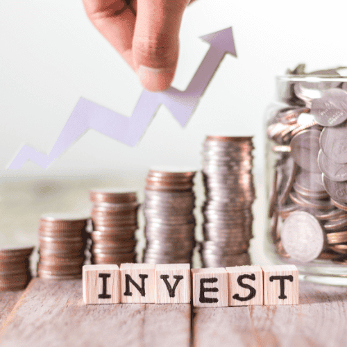Ein dynamisches Finanzbild zeigt gestapelte Münzen in aufsteigender Höhe mit einem klaren Glasgefäß voller Münzen daneben. Im Vordergrund sind Holzblöcke mit dem Wort 'INVEST' darauf positioniert. Über allem schwebt eine Hand, die einen aufsteigenden Pfeil hält, was Wachstum und positive Entwicklung symbolisiert. Der Fokus auf die Münzen und das Wort 'Invest' betont das Thema Geldanlage und die Bedeutung von Investitionen. Das Bild vermittelt Optimismus und strategische Finanzplanung.