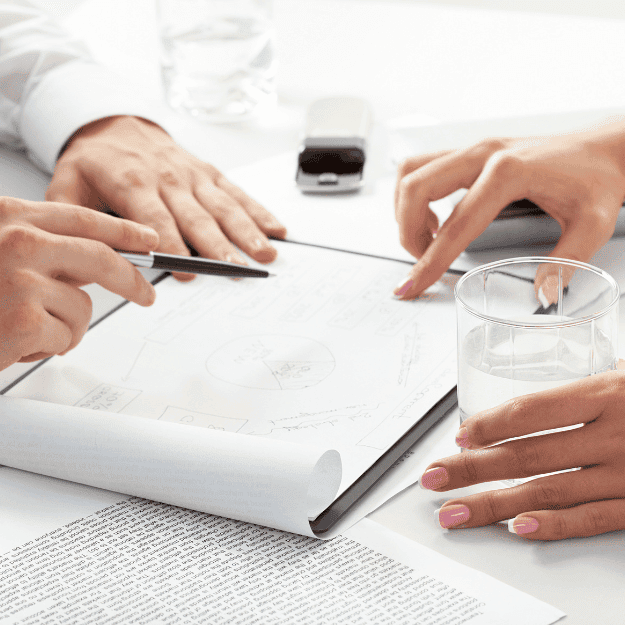 Zwei Personen diskutieren ein Finanzdokument, das auf einem Tisch ausgebreitet ist. Eine Hand mit rosa lackierten Nägeln hält ein Glas Wasser, während die andere Hand mit einem Stift auf einen wichtigen Abschnitt im Dokument zeigt. Ein Smartphone und ein Stempel sind auch auf dem Tisch sichtbar, was auf eine professionelle Beratungssituation hinweist.