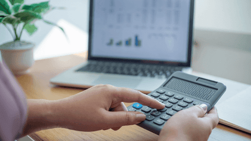 Detailansicht einer Person, die einen Taschenrechner benutzt, um Finanzdaten auf einem Laptop zu analysieren, betont die Wichtigkeit von präzisem Cashflow-Management und finanzieller Planung in einem gut beleuchteten Büro.