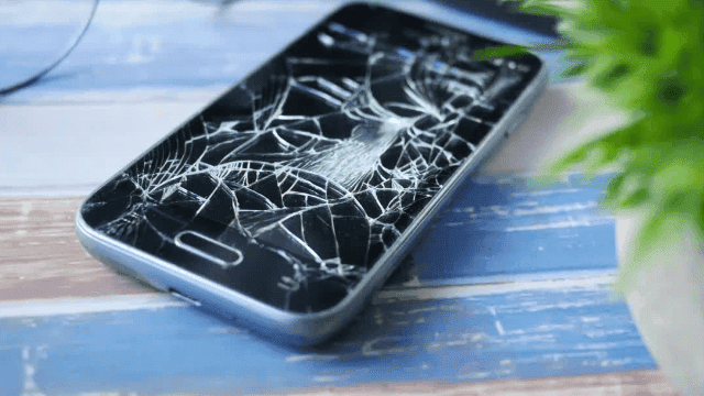 Zersplittertes Smartphone-Display liegt auf einem blauen Holztisch neben einer Pflanze, symbolisiert die Notwendigkeit einer Haftpflichtversicherung für mobile Geräte.