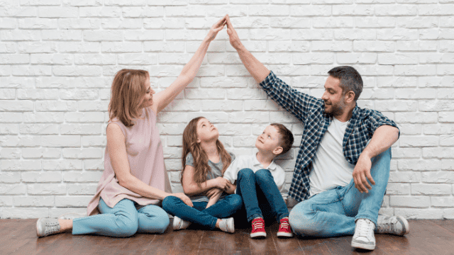 Glückliche Familie mit zwei Kindern macht zu Hause ein High-Five vor einer weißen Backsteinwand, symbolisiert Familienzusammenhalt und finanzielle Sicherheit.