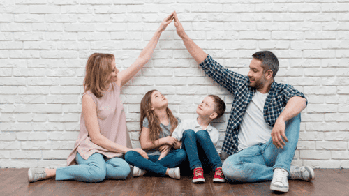 Glückliche Familie mit zwei Kindern macht zu Hause ein High-Five vor einer weißen Backsteinwand, symbolisiert Familienzusammenhalt und finanzielle Sicherheit.