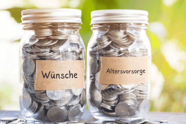 Zwei Glas-Sparschweine gefüllt mit Münzen, beschriftet mit 'Wünsche' und 'Altersvorsorge' auf einem sonnigen Tisch, symbolisieren die Wichtigkeit der finanziellen Planung und das Sparen für kurzfristige Wünsche und langfristige Ziele wie die Rente.