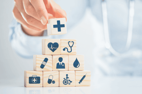 in konzeptionelles Bild, das eine Gesundheitsberatung darstellt, wobei eine Person im Arztkittel im Hintergrund unscharf zu sehen ist, während im Vordergrund Holzwürfel mit verschiedenen Gesundheits- und Medizinsymbolen wie einem Herz, einem Tropfen, einem Rollstuhl und einem Stethoskop sorgfältig gestapelt werden. Dies symbolisiert die durchdachte Planung und Absicherung im Gesundheitswesen, die eine private Krankenversicherungsberatung bietet.