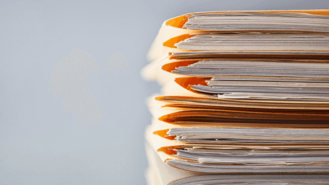 Das Bild zeigt eine Beratungssituation, in der eine Person ein Dokument über den Tisch zu einer anderen Person reicht, die erwartungsvoll die Hände verschränkt hält. Im Vordergrund ist ein kleines Modellhaus auf dem Tisch platziert, was den Fokus auf Immobilieninvestitionen und Beratungsdienstleistungen legt. Die Szene spielt sich in einem hellen Büro ab und vermittelt ein professionelles, kundenorientiertes Beratungsgespräch. Ideal für eine Webseite, die Dienstleistungen rund um Immobilien und Finanzberatung anbietet.