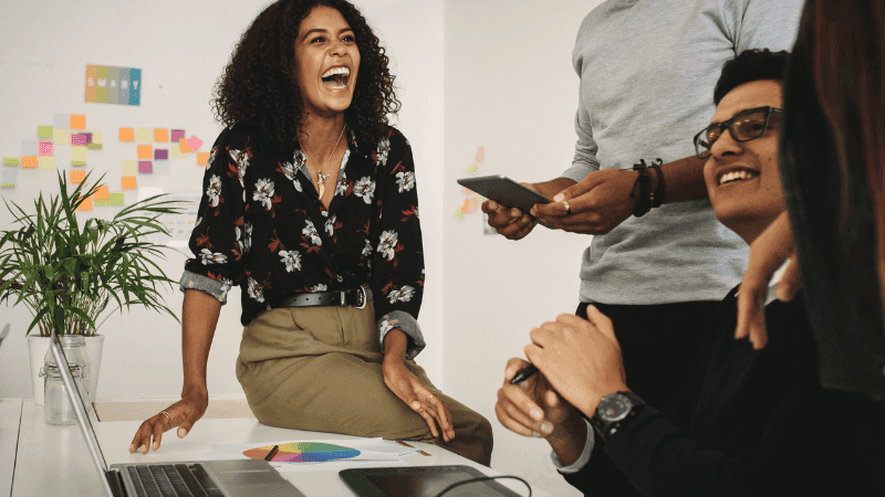 Fröhliches Brainstorming-Team in einem kreativen Büro, lachende Frau mittleren Alters im Mittelpunkt während einer entspannten Besprechung, umgeben von Kollegen mit moderner Technologie und lebhaften Diskussionen über innovative Finanzstrategien.