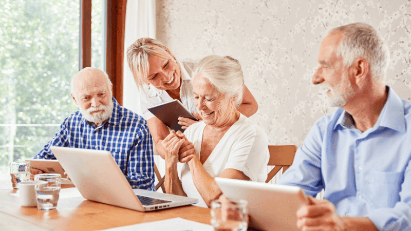 Senior*innen lernen im interaktiven Workshop 'My First Million Game', finanzielle Konzepte und Investitionen zu verstehen, unterstützt von einer freundlichen Betreuerin, die Tablet und Laptop nutzt, um Altersvorsorge und Geldanlage spielerisch zu vermitteln.