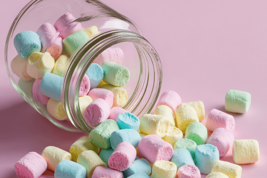 Glas mit bunten Marshmallows vor rosa Hintergrund – Symbolbild für den Marshmallow-Test, Belohnungsaufschub, Finanzpsychologie und langfristiges Denken bei Geldentscheidungen.