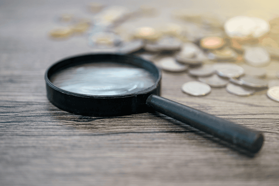 Lupe über Münzen als Symbol für transparente Kostenanalyse, unabhängigen Finanzvergleich und ganzheitliche Finanzplanung mit Fokus auf Preis-Leistungs-Verhältnis statt isolierter Kostenbetrachtung.